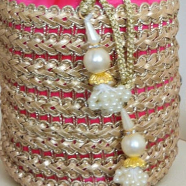 A decorative cylindrical drawstring pouch or bag made of woven beige and gold material, with a bright pink fabric top, pearl-like bead tassels, and pearl hanging ornaments. It is displayed on a round wood slice stand against a draped satin backdrop, with a pink flower and a white vase of small white blossoms nearby.