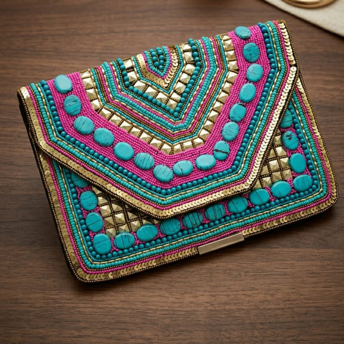 A vibrant, hand-embroidered clutch bag with a geometric mandala pattern in pink, teal, gold, and green, resting on a dark wooden surface next to a rolled cream fabric and a gold ring.