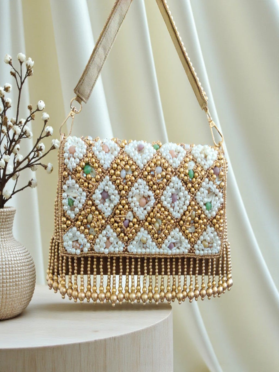 A rectangular beaded shoulder bag with a gold-tone frame and strap, decorated with white beads and multicolored sequins in a geometric diamond pattern, resting on a light wood surface with a cream draped fabric background.