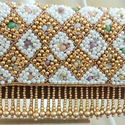 A rectangular beaded shoulder bag with a gold-tone frame and strap, decorated with white beads and multicolored sequins in a geometric diamond pattern, resting on a light wood surface with a cream draped fabric background.