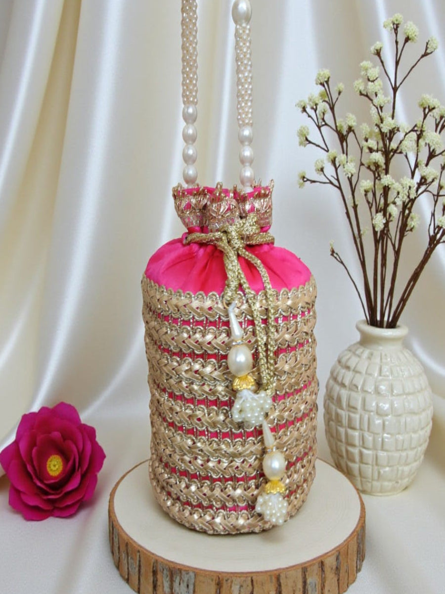 A decorative cylindrical drawstring pouch or bag made of woven beige and gold material, with a bright pink fabric top, pearl-like bead tassels, and pearl hanging ornaments. It is displayed on a round wood slice stand against a draped satin backdrop, with a pink flower and a white vase of small white blossoms nearby.