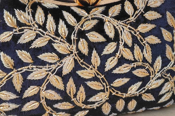 Navy blue handbag with gold leaf patterns on a wooden surface with a vase of flowers in the background.