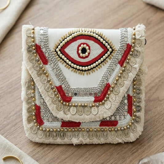 A handcrafted clutch bag with a white base, detailed with a central red and black evil eye motif, red geometric accents, and silver coin fringe, displayed on a wooden tabletop with neutral props.